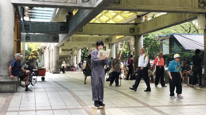 An artist standing at an open public space in the middle of a crowd, reading a book as a performance